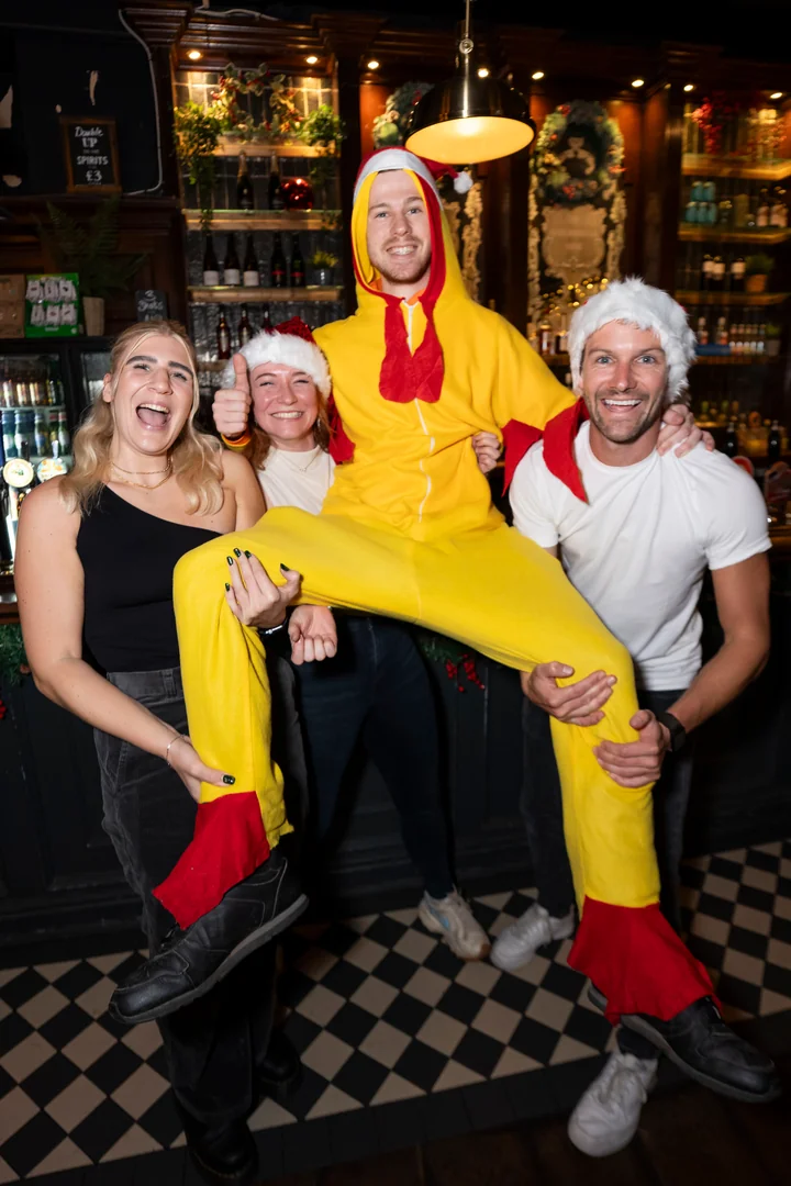 Three players in Santa hats lifting the Chicken off the ground in a festive pub